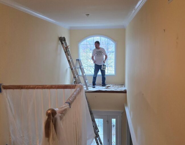 A person stands on a ladder with a paint can near a window.