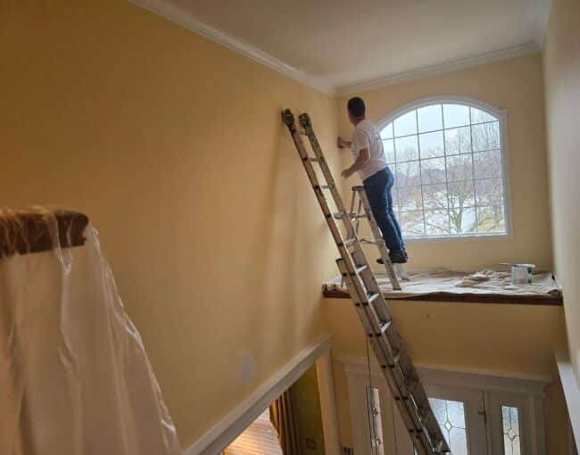 A professional painter working on an interior wall with a ladder near a window.
